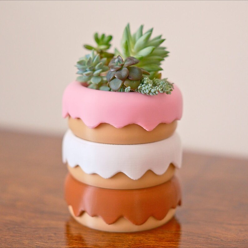 Puede incluir: Un conjunto de cuatro macetas con forma de donut en rosa, marr&oacute;n, blanco y marr&oacute;n. La maceta superior est&aacute; llena de suculentas verdes.