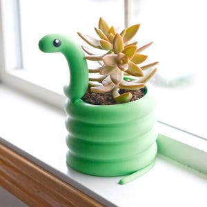 May include: A green snake-shaped planter with a succulent plant. The planter is a vibrant green with a coiled body and a snake head. The succulent has yellow and green leaves. The planter is on a white windowsill.