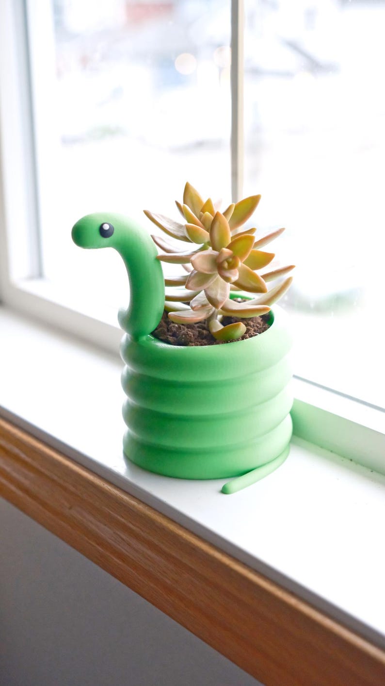 May include: A green snake-shaped planter with a succulent plant. The planter is a vibrant green with a coiled body and a snake head. The succulent has yellow and green leaves. The planter is on a white windowsill.