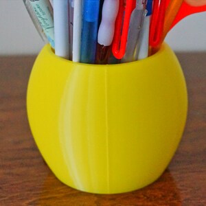 May include: A yellow ceramic pencil holder with a smooth, rounded shape. It is filled with various pens, pencils, and a pair of scissors.