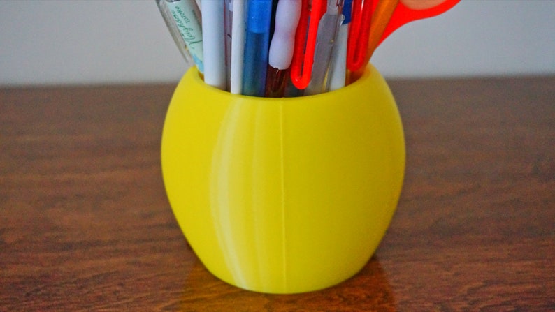 May include: A yellow ceramic pencil holder with a smooth, rounded shape. It is filled with various pens, pencils, and a pair of scissors.