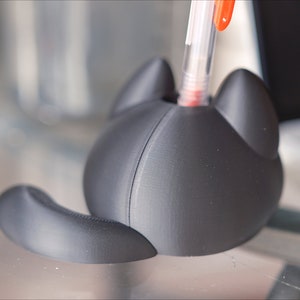 Op de afbeelding: Een zwarte 3D-geprinte penhouder in de vorm van een kat met een pen die in de bovenkant is gestoken. De houder heeft een rond lichaam en een staart.
