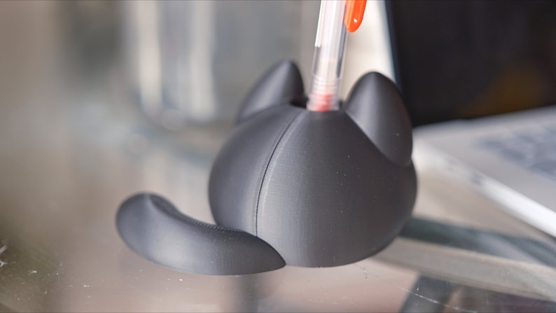 Op de afbeelding: Een zwarte 3D-geprinte penhouder in de vorm van een kat met een pen die in de bovenkant is gestoken. De houder heeft een rond lichaam en een staart.