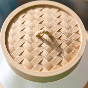 Puede incluir: Una tapa de madera con un patr&oacute;n tejido y un asa. La tapa es beige y tiene una veta de madera clara.