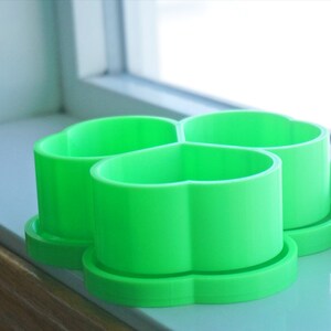 May include: Three bright green, heart-shaped cookie cutters sit on a windowsill. Each cutter has a round base and a cylindrical body with a scalloped top. The cutters are made of plastic and are arranged close together.
