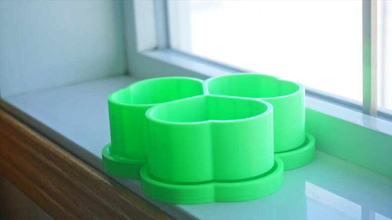 May include: Three bright green, heart-shaped cookie cutters sit on a windowsill. Each cutter has a round base and a cylindrical body with a scalloped top. The cutters are made of plastic and are arranged close together.