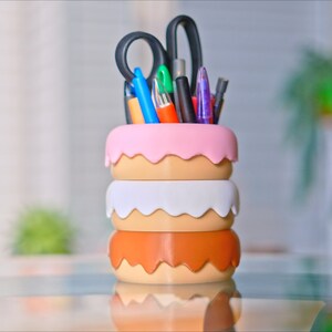 Puede incluir: Un organizador de escritorio de tres niveles con forma de donut en rosa, blanco y marr&oacute;n con una variedad de bol&iacute;grafos, l&aacute;pices y tijeras que sobresalen de la parte superior.