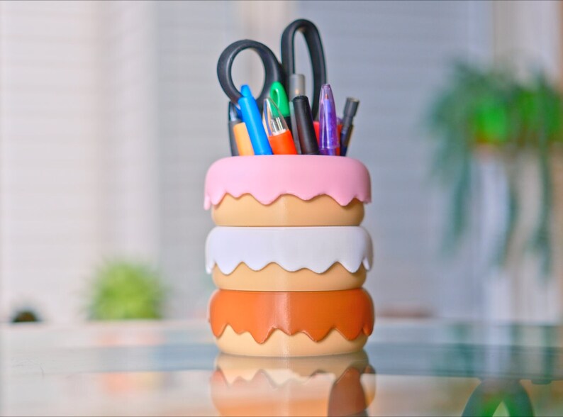Puede incluir: Un organizador de escritorio de tres niveles con forma de donut en rosa, blanco y marr&oacute;n con una variedad de bol&iacute;grafos, l&aacute;pices y tijeras que sobresalen de la parte superior.