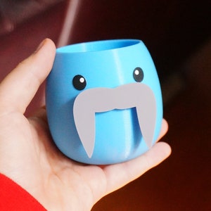 May include: A light blue ceramic planter shaped like a walrus with a gray mustache. The planter has two black eyes and a white mouth.
