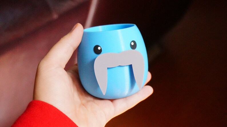 May include: A light blue ceramic planter shaped like a walrus with a gray mustache. The planter has two black eyes and a white mouth.