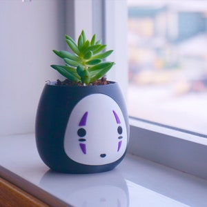May include: A small, black ceramic pot with a white face design and purple accents, holding a green succulent plant. The pot is oval-shaped and sits on a white windowsill. The background is blurred, showing a window and a snowy outdoor scene.