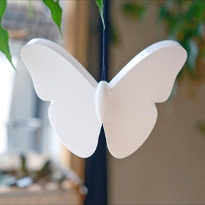 May include: A white butterfly decoration suspended by a thin black string. The butterfly has a symmetrical design with rounded wings and a central body. Green leaves and a blurred background are visible.
