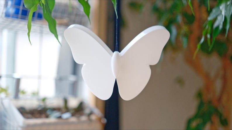 May include: A white butterfly decoration suspended by a thin black string. The butterfly has a symmetrical design with rounded wings and a central body. Green leaves and a blurred background are visible.