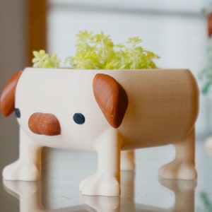 May include: A white ceramic planter shaped like a dog with brown ears and nose. The planter has four legs and is holding a small green plant.