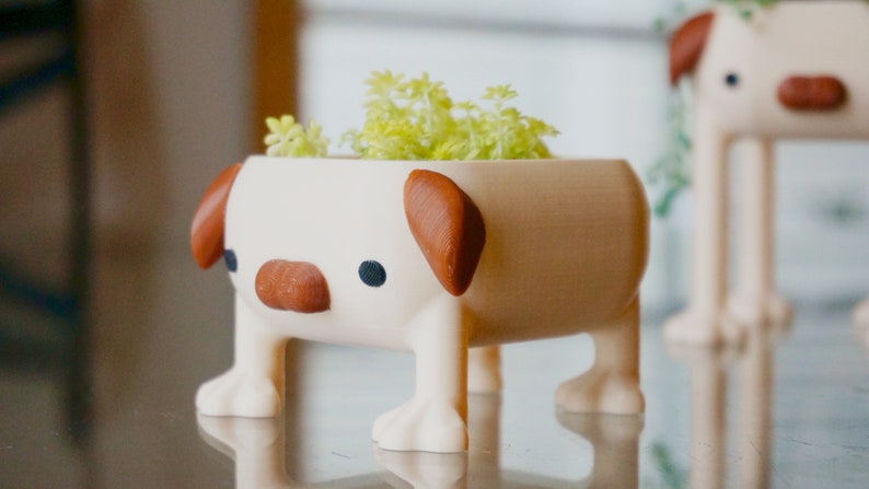 May include: A white ceramic planter shaped like a dog with brown ears and nose. The planter has four legs and is holding a small green plant.