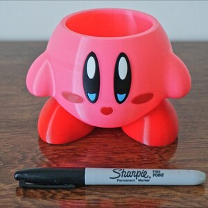 Puede incluir: Maceta rosa de impresi&oacute;n 3D con forma del personaje de videojuego Kirby. La maceta tiene una peque&ntilde;a abertura en la parte superior para plantar. La maceta est&aacute; sobre una superficie de madera.