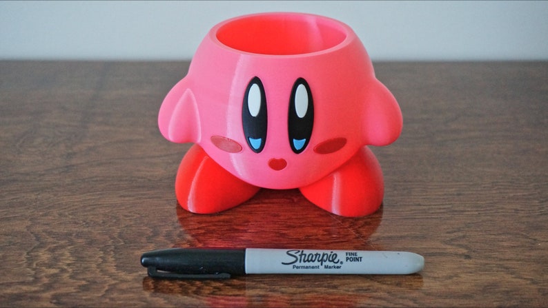 Puede incluir: Maceta rosa de impresi&oacute;n 3D con forma del personaje de videojuego Kirby. La maceta tiene una peque&ntilde;a abertura en la parte superior para plantar. La maceta est&aacute; sobre una superficie de madera.