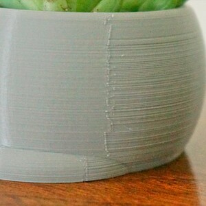 Pode incluir: Um vaso de planta cinza claro impresso em 3D com uma superf&iacute;cie lisa e arredondada. O vaso &eacute; feito com um design texturizado em camadas.