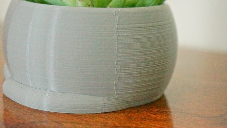 Pode incluir: Um vaso de planta cinza claro impresso em 3D com uma superf&iacute;cie lisa e arredondada. O vaso &eacute; feito com um design texturizado em camadas.