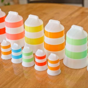 Może przedstawiać: Kolekcja białych, plastikowych przedmiot&oacute;w w kształcie butelek z kolorowymi poziomymi paskami w r&oacute;żnych rozmiarach. Paski są w kolorze pomarańczowym, r&oacute;żowym, ż&oacute;łtym, zielonym i niebieskim. Przedmioty ułożone są na drewnianej powierzchni.