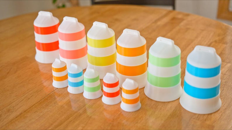 Może przedstawiać: Kolekcja białych, plastikowych przedmiot&oacute;w w kształcie butelek z kolorowymi poziomymi paskami w r&oacute;żnych rozmiarach. Paski są w kolorze pomarańczowym, r&oacute;żowym, ż&oacute;łtym, zielonym i niebieskim. Przedmioty ułożone są na drewnianej powierzchni.