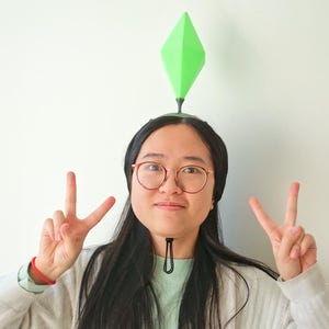 May include: A person wearing a green diamond-shaped plumbob headpiece, a symbol from The Sims video game, with a black headband. The person is wearing glasses and making a peace sign with both hands.