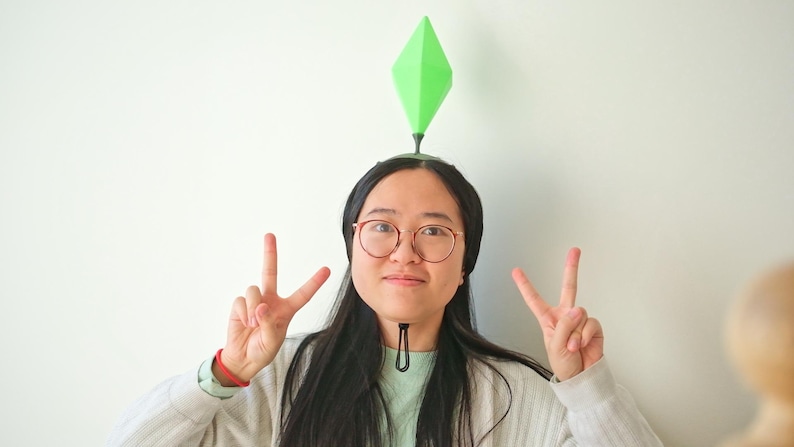 May include: A person wearing a green diamond-shaped plumbob headpiece, a symbol from The Sims video game, with a black headband. The person is wearing glasses and making a peace sign with both hands.