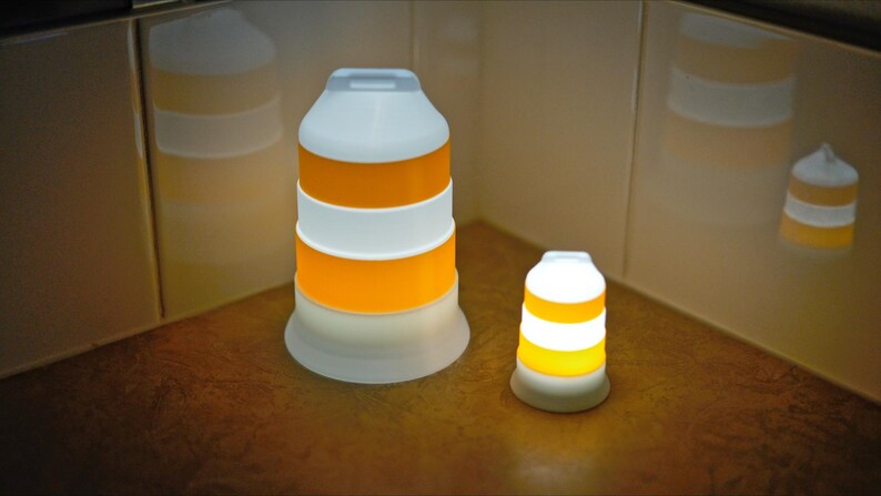 Może przedstawiać: Dwie cylindryczne, układane w stosy lampy LED. Większa lampa jest biało-pomarańczowa, z białą g&oacute;rą i podstawą. Mniejsza lampa ma podobny design. Obie lampy są podświetlone, emitując ciepłą poświatę. Lampy znajdują się na brązowej powierzchni.