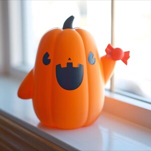 May include: A smiling orange pumpkin figurine with a black face and a candy cane in its hand. The pumpkin is sitting on a white surface.