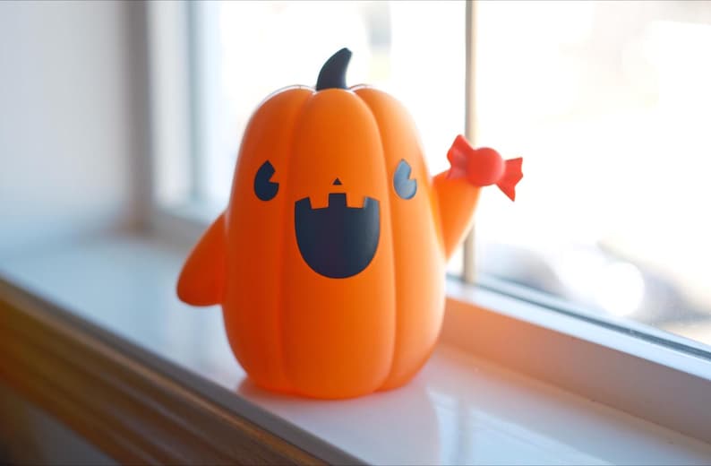 May include: A smiling orange pumpkin figurine with a black face and a candy cane in its hand. The pumpkin is sitting on a white surface.