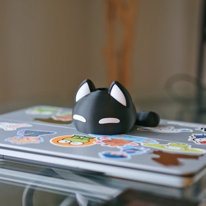 Op de afbeelding: Een zwarte, katvormige telefoonstandaard met witte oren en ogen, die op een laptop staat die bedekt is met kleurrijke stickers.