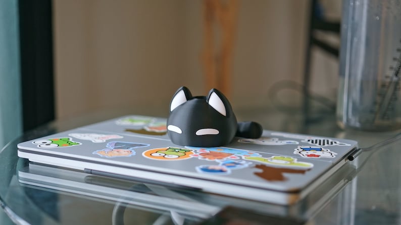 Op de afbeelding: Een zwarte, katvormige telefoonstandaard met witte oren en ogen, die op een laptop staat die bedekt is met kleurrijke stickers.