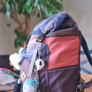 May include: A multi-colored backpack with a blue top, red center, and brown bottom. The backpack has a front pocket and is decorated with several small plush keychains. The backpack is sitting on a wooden table.