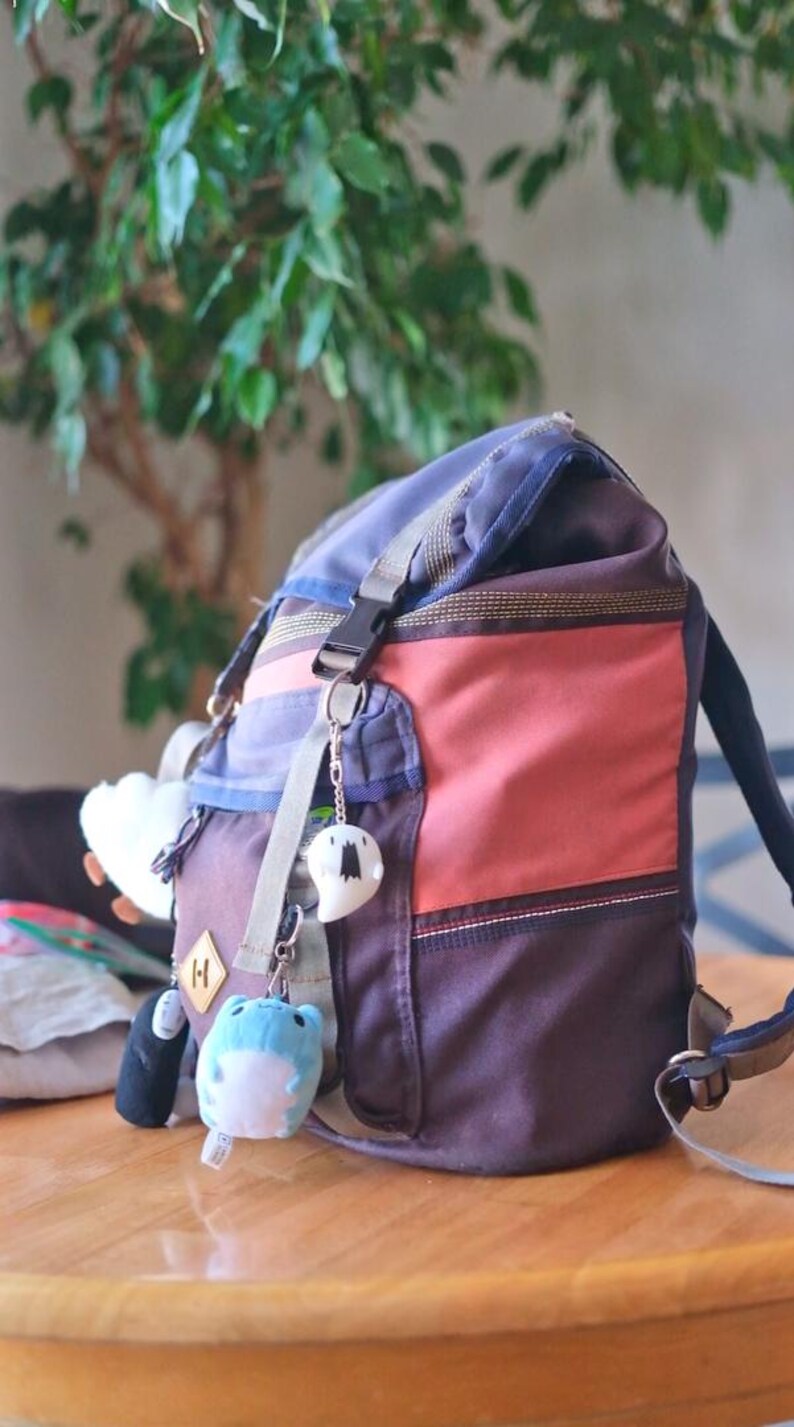 May include: A multi-colored backpack with a blue top, red center, and brown bottom. The backpack has a front pocket and is decorated with several small plush keychains. The backpack is sitting on a wooden table.