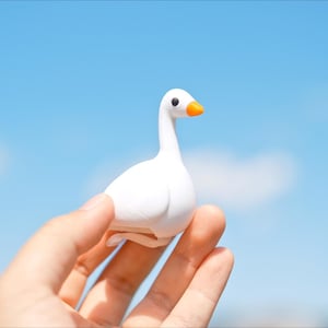 May include: A small, white goose figurine with an orange beak and black eyes is held against a bright blue sky. The goose is the primary focus, showcasing its smooth, rounded form. The background is a clear, sunny sky.