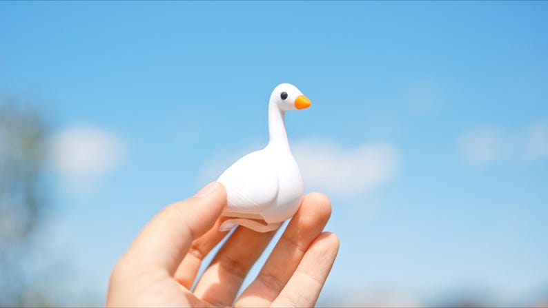 May include: A small, white goose figurine with an orange beak and black eyes is held against a bright blue sky. The goose is the primary focus, showcasing its smooth, rounded form. The background is a clear, sunny sky.