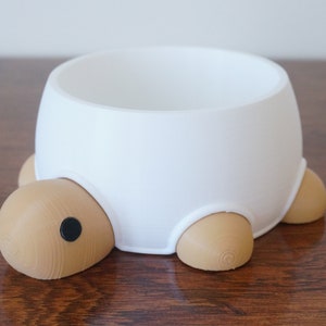 May include: White ceramic pet bowl shaped like a turtle with brown legs and a black eye. The bowl is sitting on a wooden surface.