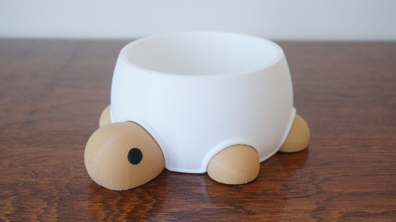 May include: White ceramic pet bowl shaped like a turtle with brown legs and a black eye. The bowl is sitting on a wooden surface.