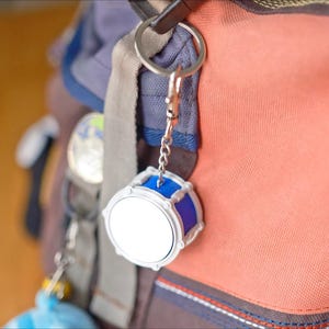May include: A silver and blue drum-shaped keychain hangs from a backpack. The keychain has a white drum face and blue accents. The backpack is a mix of orange, blue, and gray fabrics.