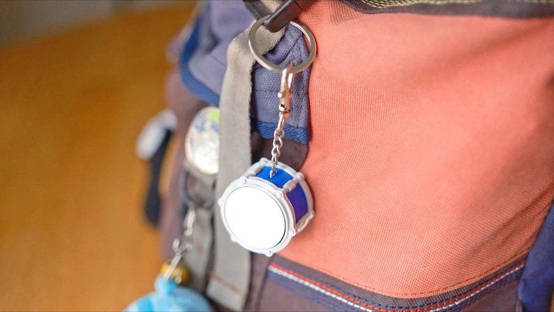 May include: A silver and blue drum-shaped keychain hangs from a backpack. The keychain has a white drum face and blue accents. The backpack is a mix of orange, blue, and gray fabrics.