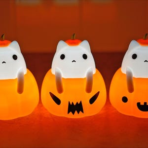 May include: Three glowing Halloween cat-in-pumpkin night lights. Each light features a white cat figure sitting inside an orange pumpkin with a carved face. The pumpkins have different expressions, including a jack-o'-lantern face and a smiling face.