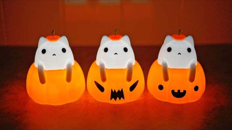 May include: Three glowing Halloween cat-in-pumpkin night lights. Each light features a white cat figure sitting inside an orange pumpkin with a carved face. The pumpkins have different expressions, including a jack-o'-lantern face and a smiling face.