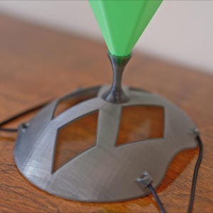 May include: A close-up of a modern lamp base with a geometric design. The base is gray with diamond-shaped cutouts and a black stem supporting a green, cone-shaped top. Black wires extend from the base.