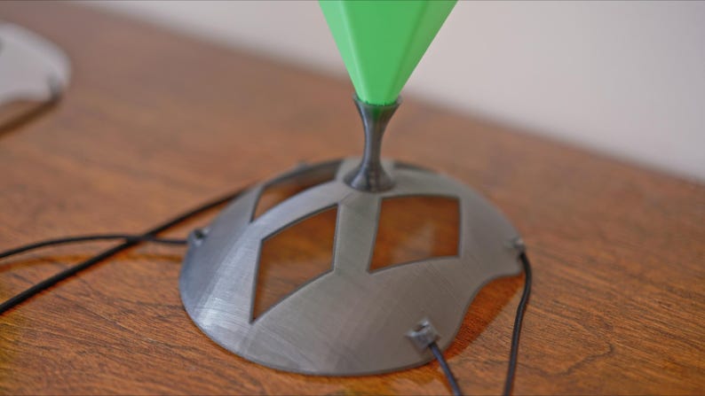 May include: A close-up of a modern lamp base with a geometric design. The base is gray with diamond-shaped cutouts and a black stem supporting a green, cone-shaped top. Black wires extend from the base.