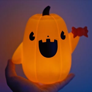 May include: A glowing orange pumpkin-shaped light with a friendly face and a candy cane in its hand. The light is held in a person's hand.