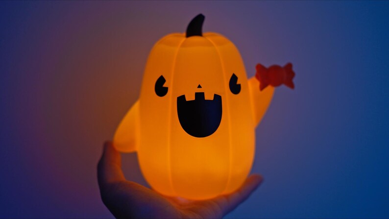 May include: A glowing orange pumpkin-shaped light with a friendly face and a candy cane in its hand. The light is held in a person's hand.