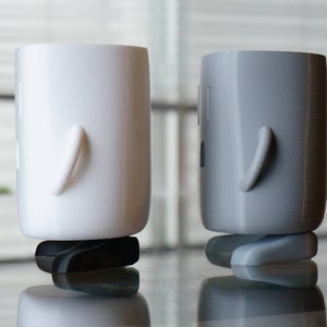 Op de afbeelding: Twee 3D-geprinte figuren, een witte en een grijze, staan ​​naast elkaar op een glazen tafel. Beide figuren hebben een rond, cilindrisch lichaam en een enkele arm die naar de zijkant uitsteekt.