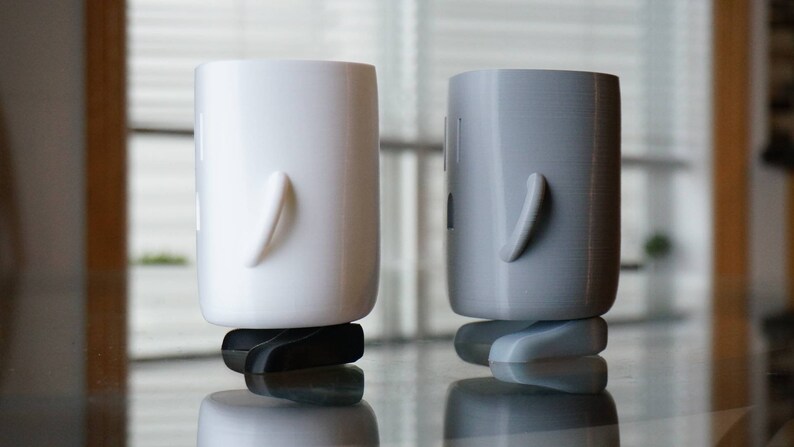 Op de afbeelding: Twee 3D-geprinte figuren, een witte en een grijze, staan ​​naast elkaar op een glazen tafel. Beide figuren hebben een rond, cilindrisch lichaam en een enkele arm die naar de zijkant uitsteekt.