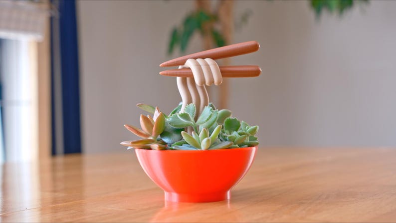 May include: A whimsical succulent planter shaped like a ramen bowl. The bright orange bowl holds green succulents, with faux noodles and chopsticks. A unique decorative piece for home or office.