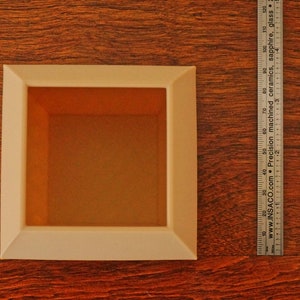 May include: A square, off-white shadow box with an orange interior sits on a wood-grain surface. A metal ruler is positioned next to the box. The shadow box is approximately 4 inches wide.